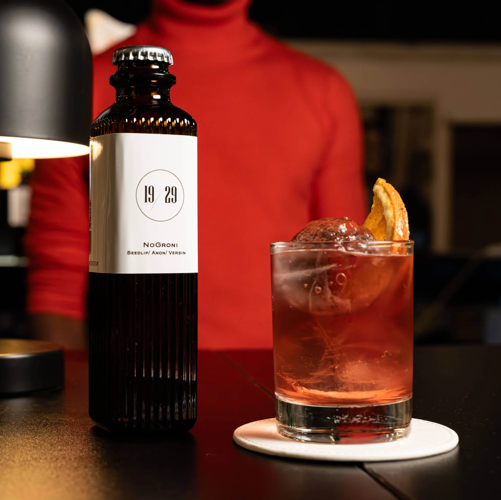 A dark brown glass bottle labeled "19 29 NOGRONI" next to a clear glass filled with a reddish-brown cocktail, garnished with an orange slice, on a dark surface with a blurred background featuring a person in a red turtleneck.