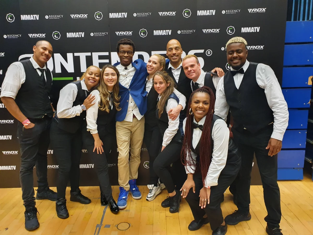 A diverse group of 10-12 professional event staff members in semi-formal attire, posing in front of a black backdrop with logos such as MMATV, REGENCY, and RDX at a hospitality event.
